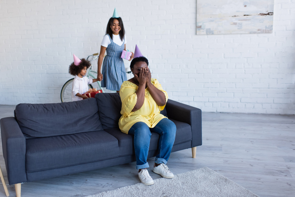 afro-amerikai anya és lánya kezében ajándékok közelében nagyi party sapka ül a kanapén, és eltakarja a szemét kézzel - Fotó, kép