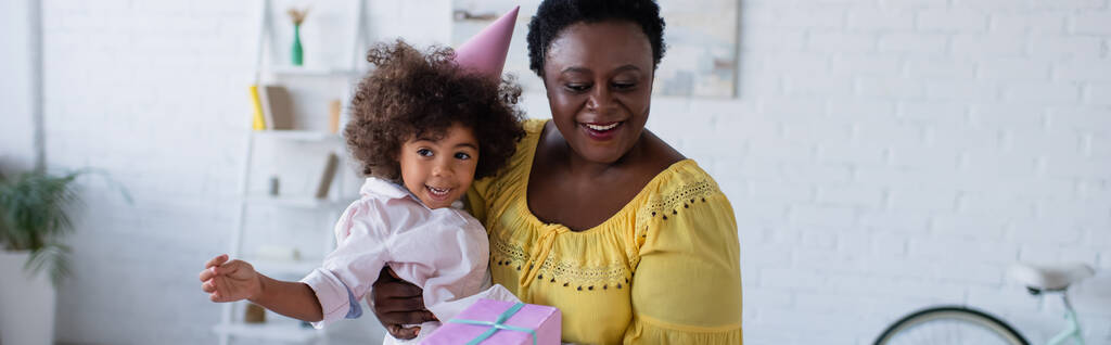 joyeuse femme afro-américaine d'âge moyen tenant sa petite-fille dans une casquette de fête et une boîte-cadeau, une bannière - Photo, image