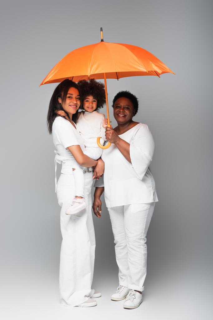 χαρούμενες αφροαμερικανικές γυναίκες και το παιδί κοιτάζοντας κάμερα κάτω από πορτοκαλί ομπρέλα σε γκρι - Φωτογραφία, εικόνα