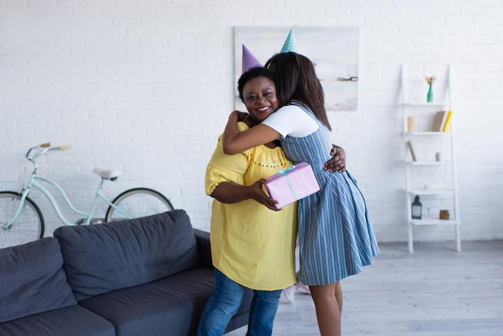 mladá africká americká žena ve straně čepice objímající šťastný matka drží dárek k narozeninám  - Fotografie, Obrázek
