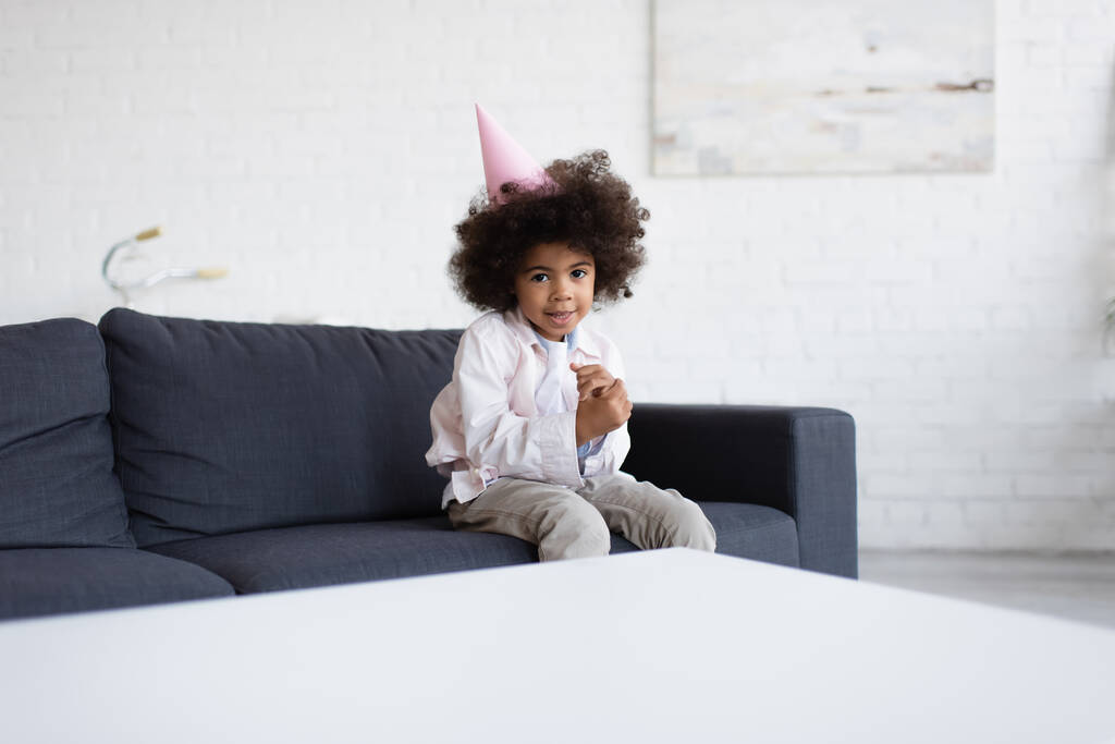 Африканская именинница в шапочке для вечеринок сидит на диване возле размытого стола и смотрит в камеру - Фото, изображение
