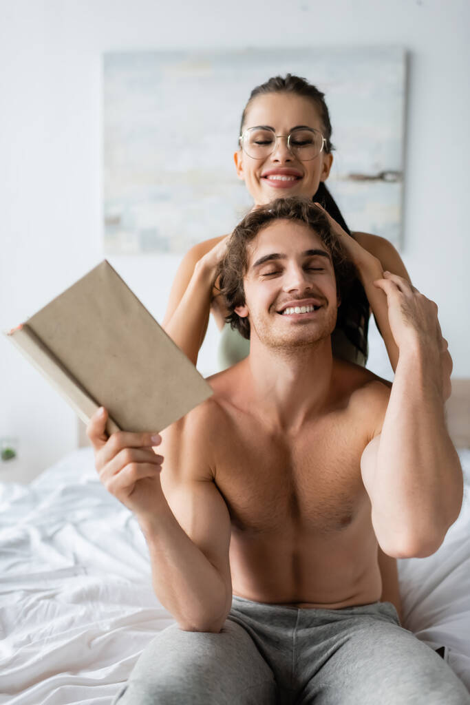 Mujer sonriente con anteojos tocando la cabeza del novio sin camisa sosteniendo libro en la cama  - Foto, imagen