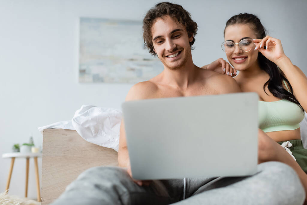 Sorrindo mulher sexy em óculos olhando para laptop borrado perto de namorado sem camisa no quarto  - Foto, Imagem