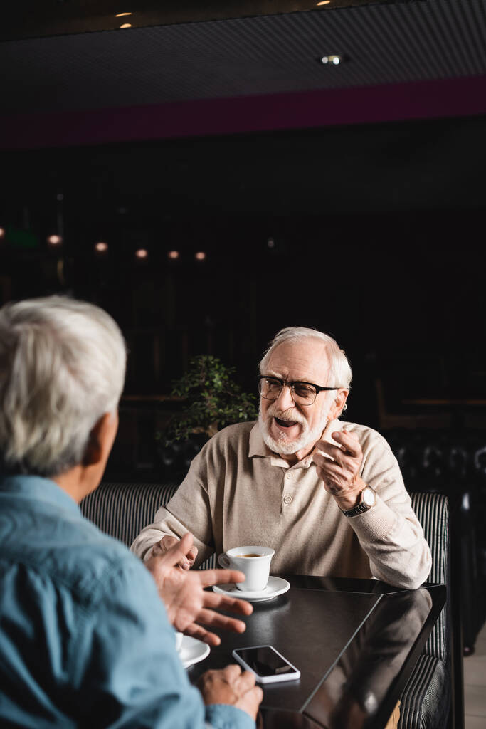 homem idoso em óculos gesticulando enquanto conversa com amigo desfocado no café - Foto, Imagem
