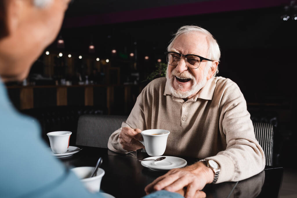 alegre hombre mayor en gafas con taza de café mientras toca la mano de un amigo borroso en la cafetería - Foto, Imagen