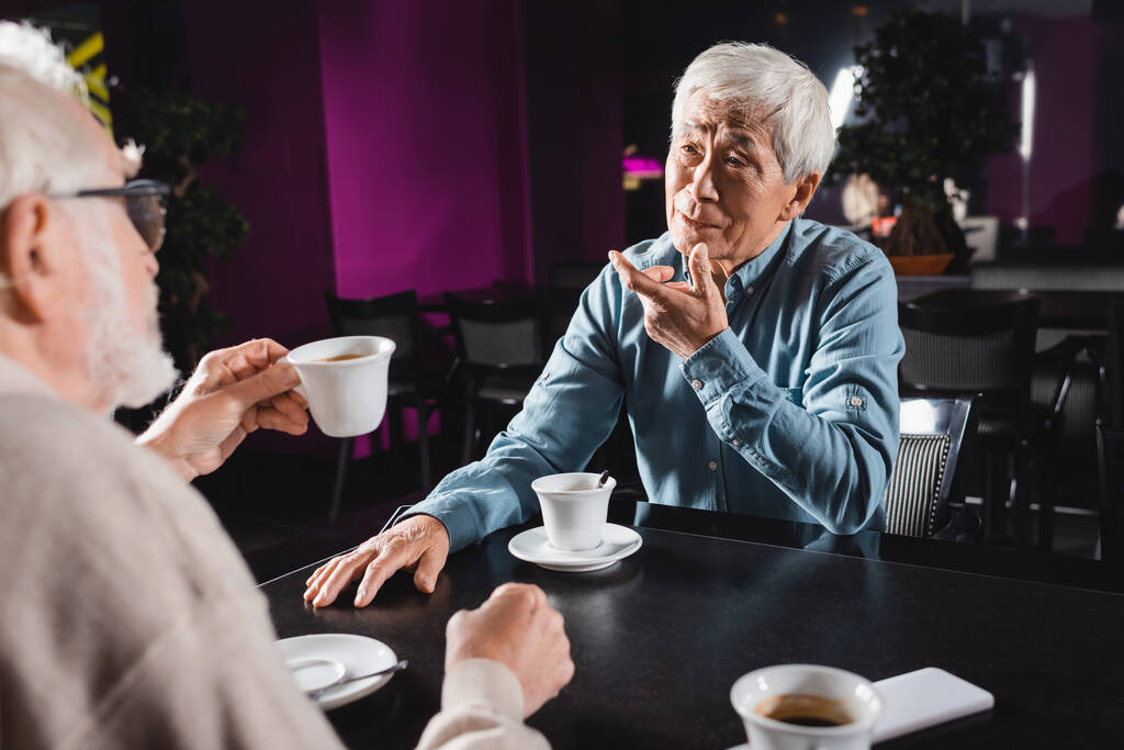 wazig man vasthouden van kopje koffie in de buurt van senior aziatische man gebaren tijdens het praten in cafe - Foto, afbeelding