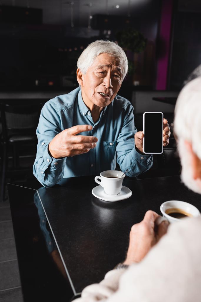 アジア系の先輩がブランクスクリーンの携帯電話を指差してカフェでぼやけた友人と時間を過ごしながら - 写真・画像