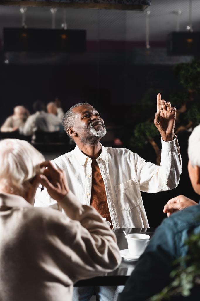 glimlachende Afrikaans-Amerikaanse man omhoog kijkend en wijzend met de vinger in de buurt van wazig senior vrienden in cafe - Foto, afbeelding