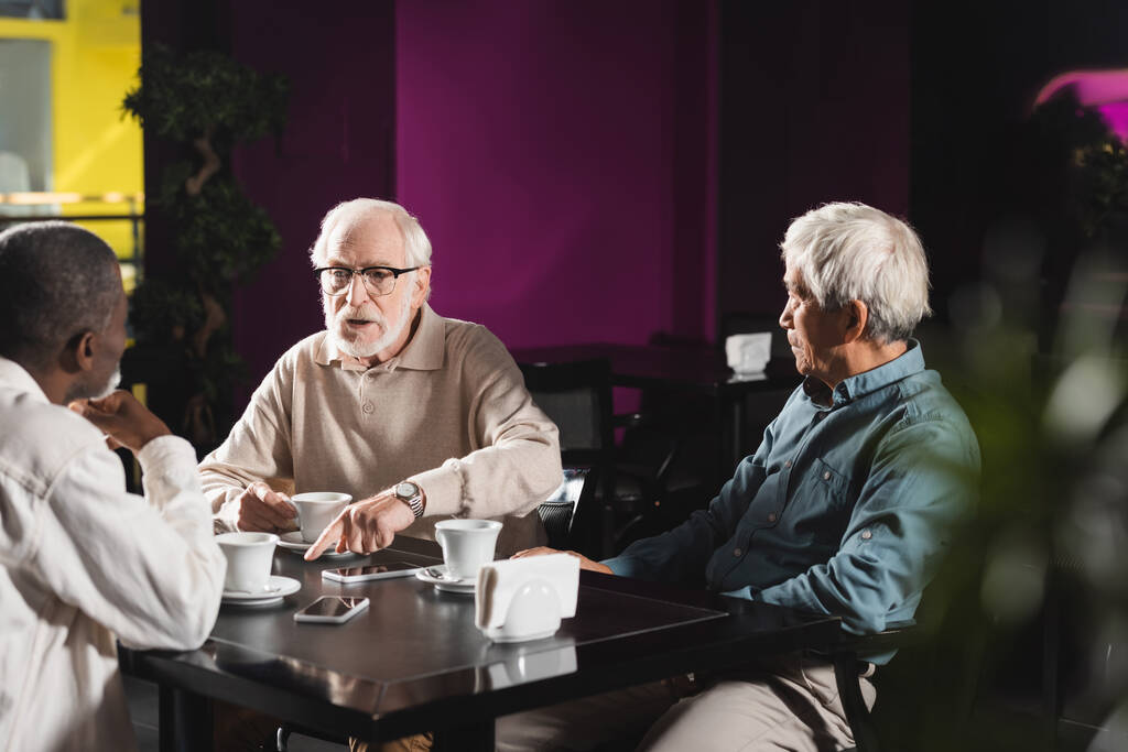 Senior mit Brille zeigt auf Smartphone mit leerem Bildschirm, während er Zeit im Café verbringt - Foto, Bild
