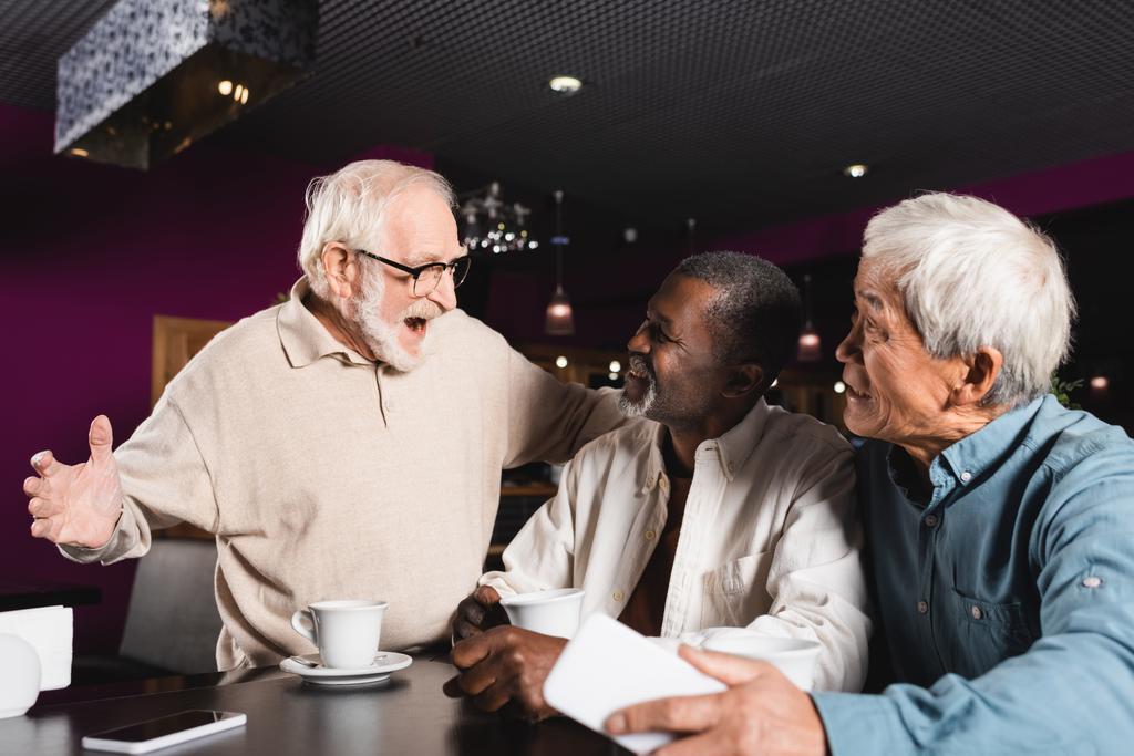 emocionado hombre mayor mostrando wow gesto cerca de felices amigos multiétnicos en la cafetería - Foto, Imagen