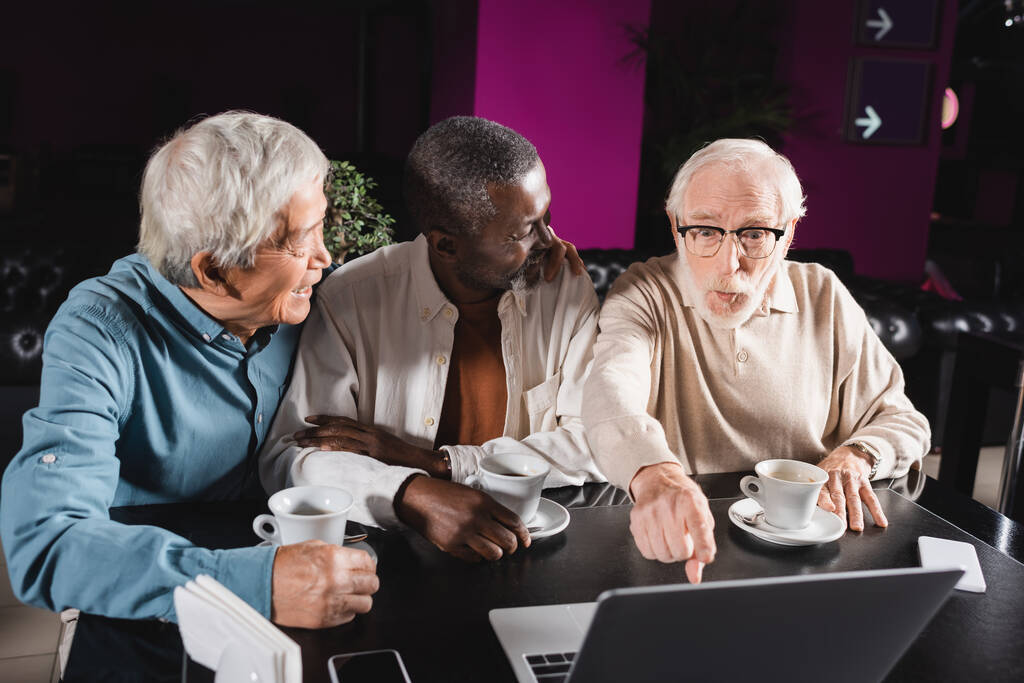 meghökkent idős férfi mutató laptop közel mosolygós multietnikus barátok kávézóban - Fotó, kép