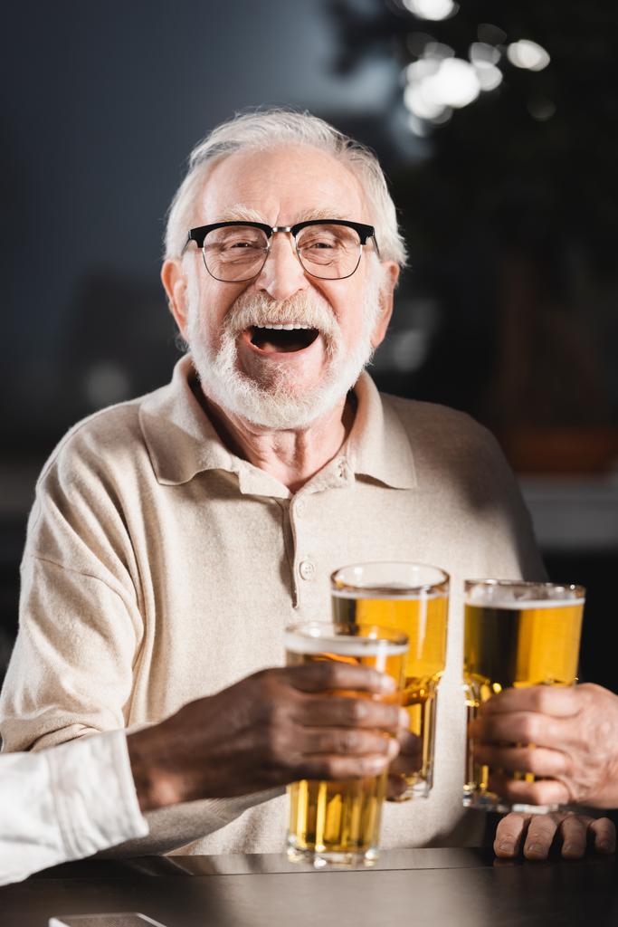 ηλικιωμένος άνδρας με γυαλιά ηλίου γελώντας ενώ έπινε ποτήρια μπύρας με θολή διαφυλετικούς φίλους - Φωτογραφία, εικόνα
