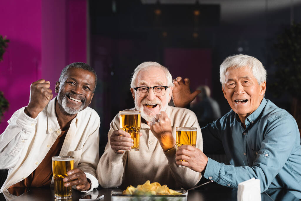 веселые старшие мультикультурные друзья с бокалами пива показывают победу жест во время просмотра футбольного матча в пабе - Фото, изображение