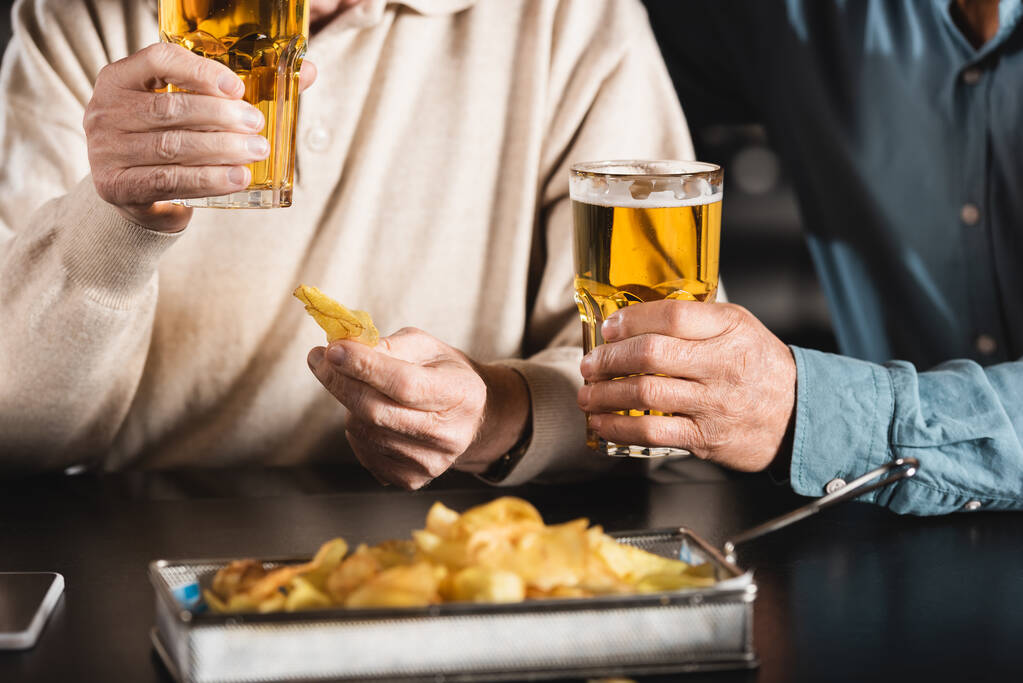 vista cortada de amigos seniores segurando copos de cerveja perto de chips borrados - Foto, Imagem