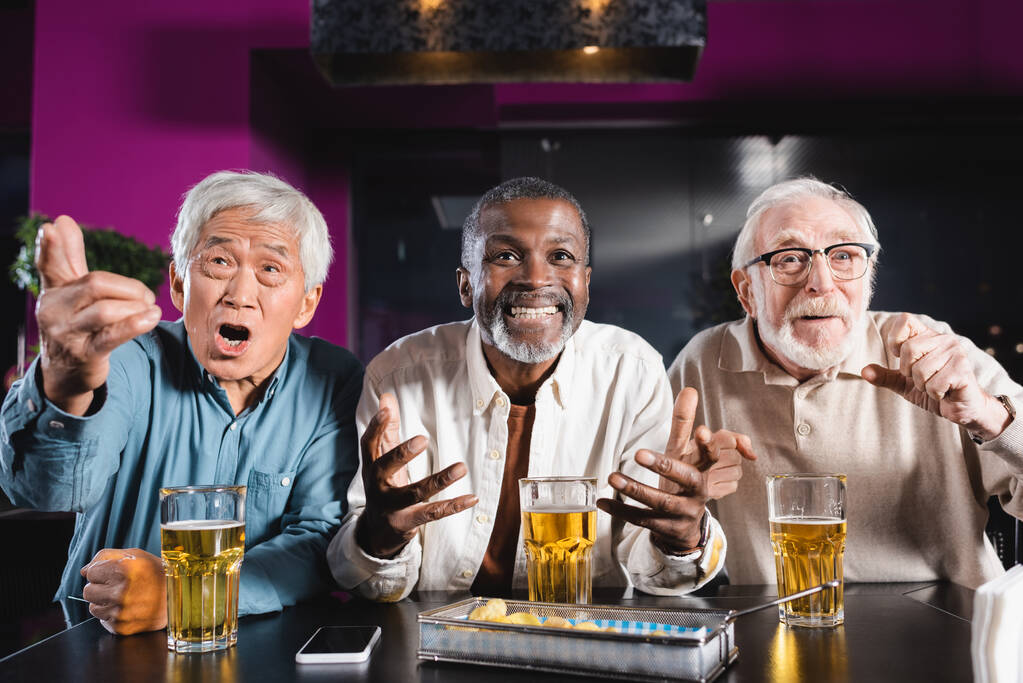 excited elderly multiethnic friends gesturing while watching soccer match in beer pub - Photo, Image