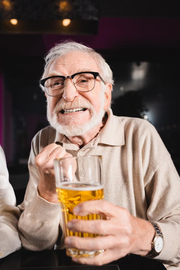 worried senior man in eyeglasses holding blurred glass of beer while watching soccer championship in pub - Photo, Image