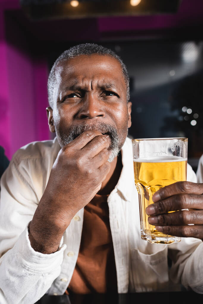 sénior afroamericano hombre sosteniendo vaso de cerveza mientras mira campeonato en bar deportivo - Foto, imagen