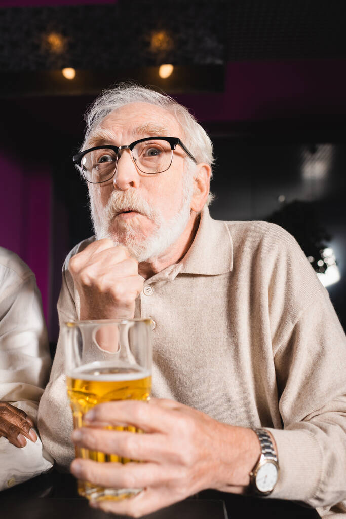 napięty senior z zaciśniętą pięścią i szklanką piwa oglądający mecz piłki nożnej w barze sportowym - Zdjęcie, obraz