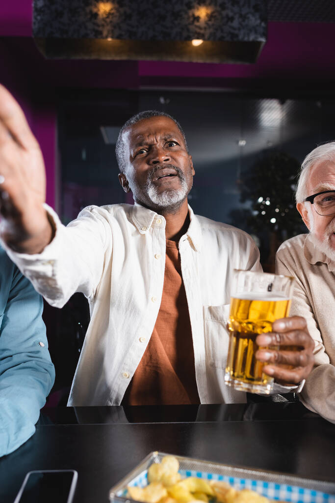 bouleversé homme afro-américain pointant avec la main tout en regardant match de football avec des amis âgés dans le pub - Photo, image