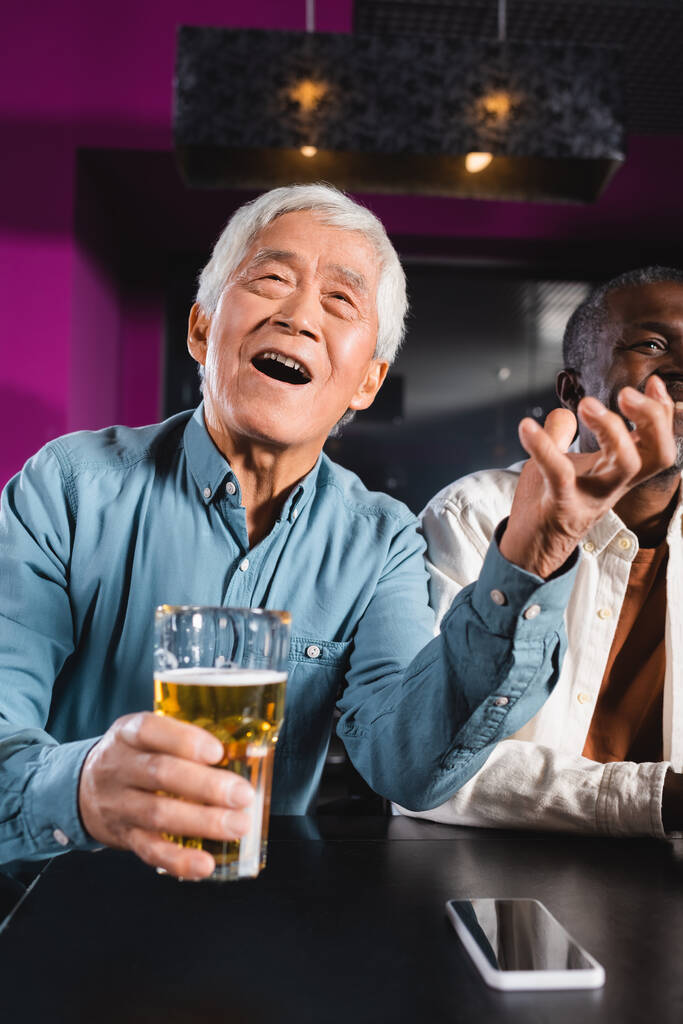 espantado asiático homem com copo de cerveja assistindo campeonato no esporte bar perto sênior multiétnico amigos - Foto, Imagem