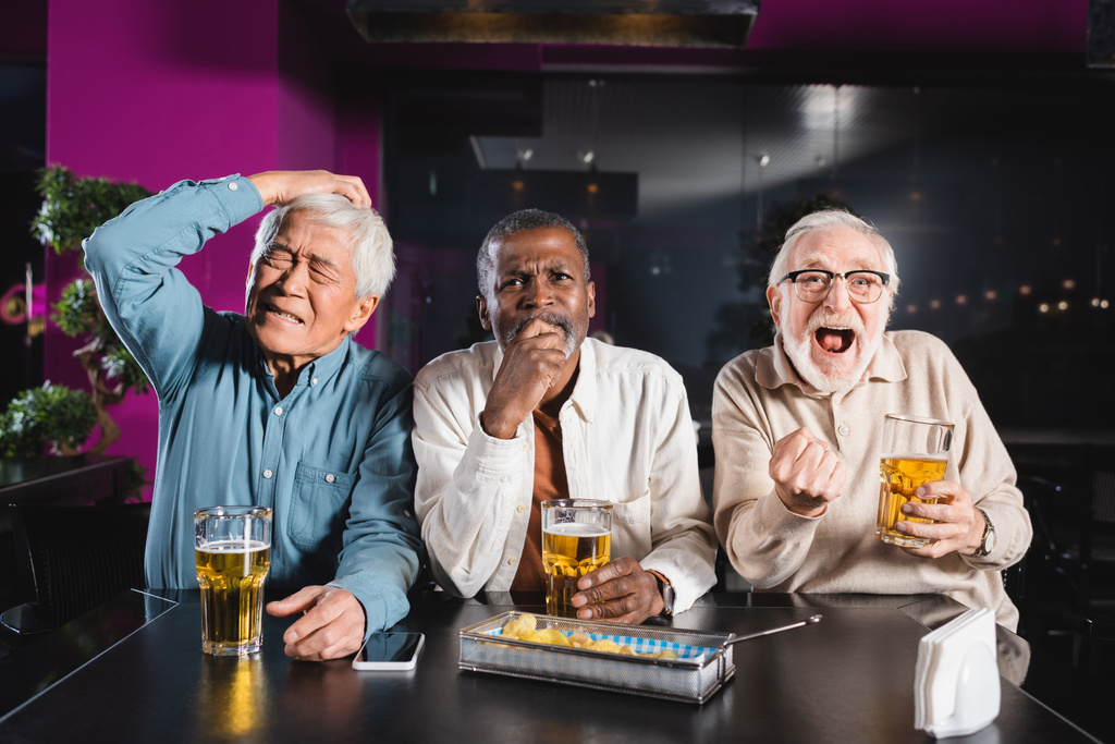 изумленный и счастливый пожилой человек, показывающий победный жест, смотря футбол с расстроенными межрасовыми друзьями в пабе - Фото, изображение