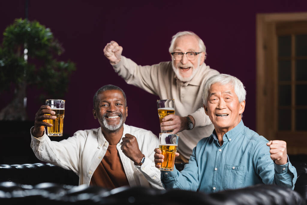 χαρούμενους πολυπολιτισμικούς φίλους με ποτήρια μπύρας που δείχνουν νικηφόρα χειρονομία σε αθλητικό μπαρ - Φωτογραφία, εικόνα