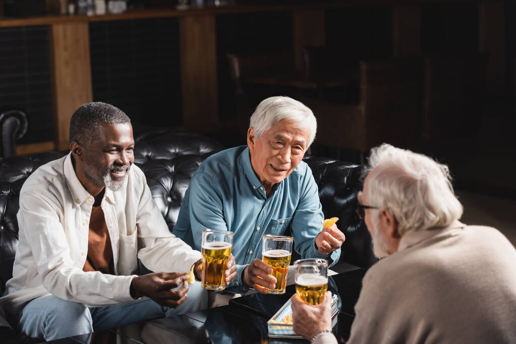 senior multicultural friends holding glasses of beer while spending time in pub - Photo, Image