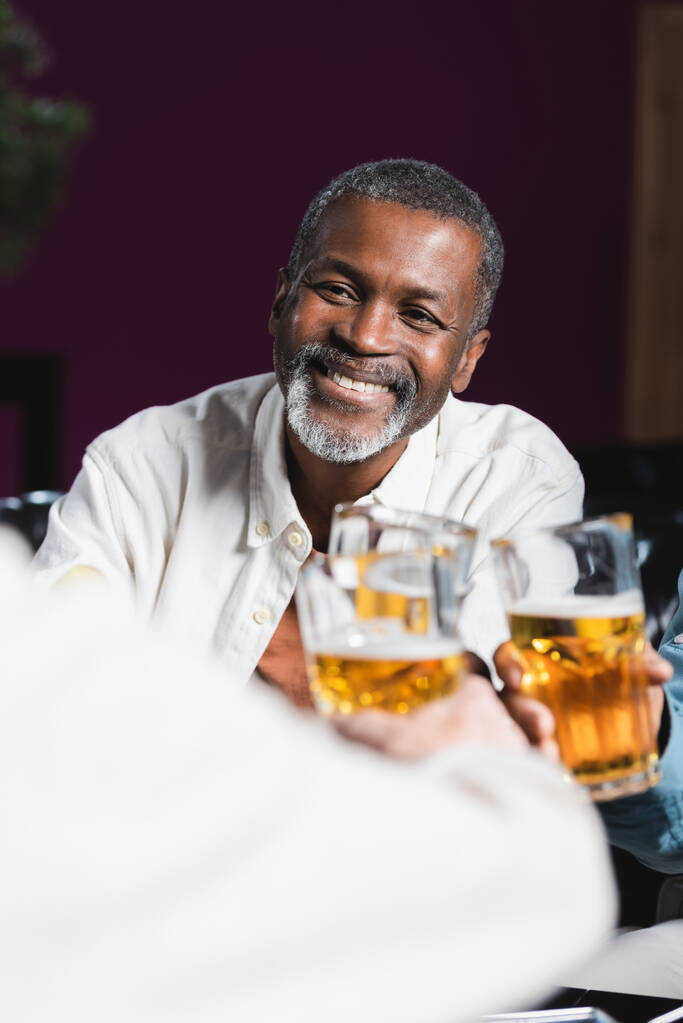alegre africano americano hombre tintineo cerveza vasos con borrosa senior amigos en pub - Foto, Imagen