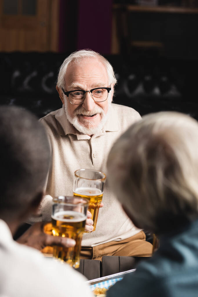старший чоловік в окулярах посміхається, тримаючи келих пива біля розмитих друзів у пабі
 - Фото, зображення