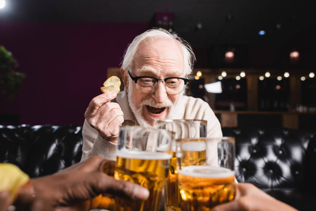 aufgeregter Senior hält Chips in der Hand, während er mit verschwommenen interrassischen Freunden Biergläser klirrt - Foto, Bild