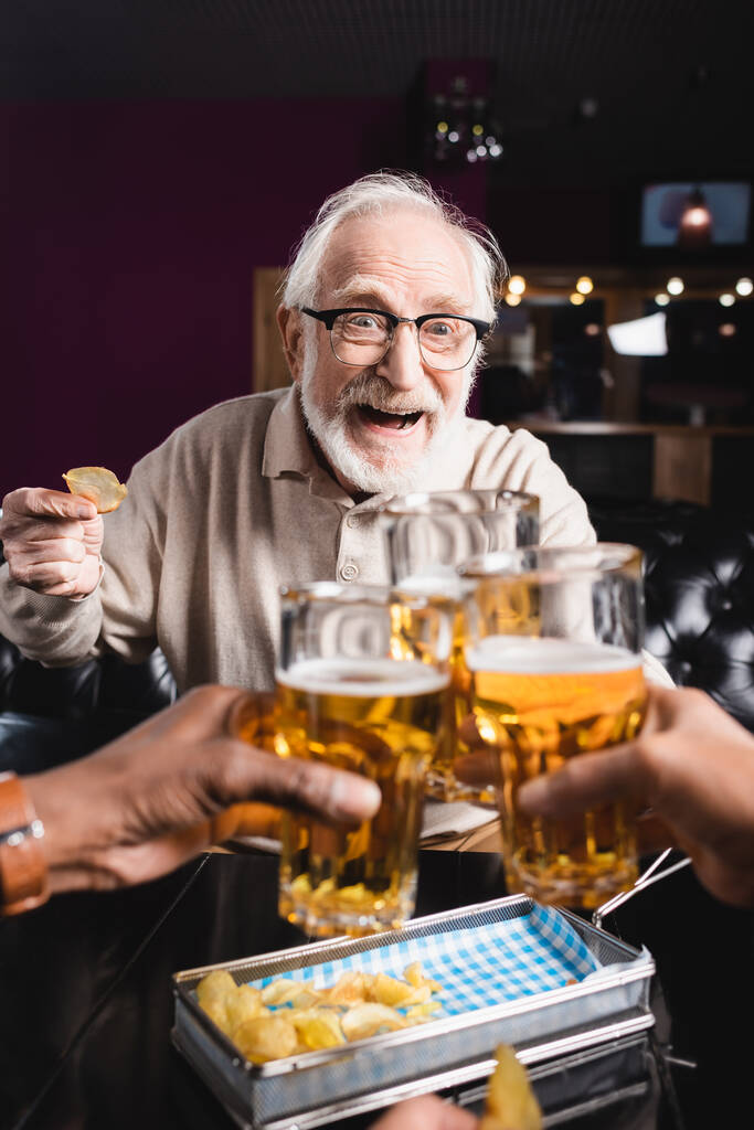 vzrušený starší muž v brýlích cinkání pivo brýle s rozmazané mezirasové přátelé - Fotografie, Obrázek