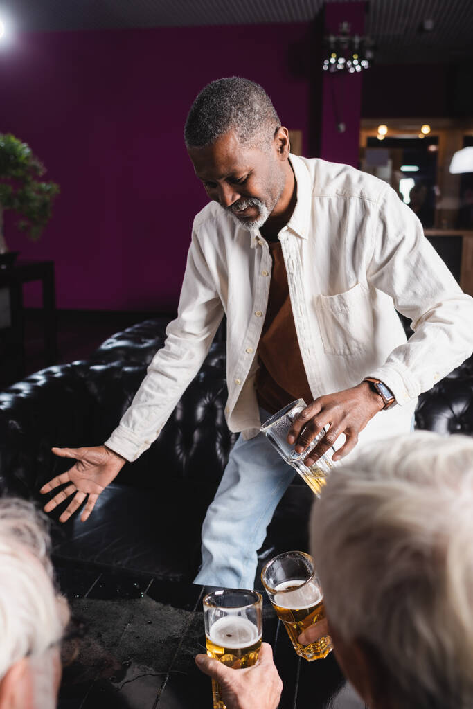 homme afro-américain senior tenant un verre de bière tout en se tenant près des amis flous dans le pub - Photo, image