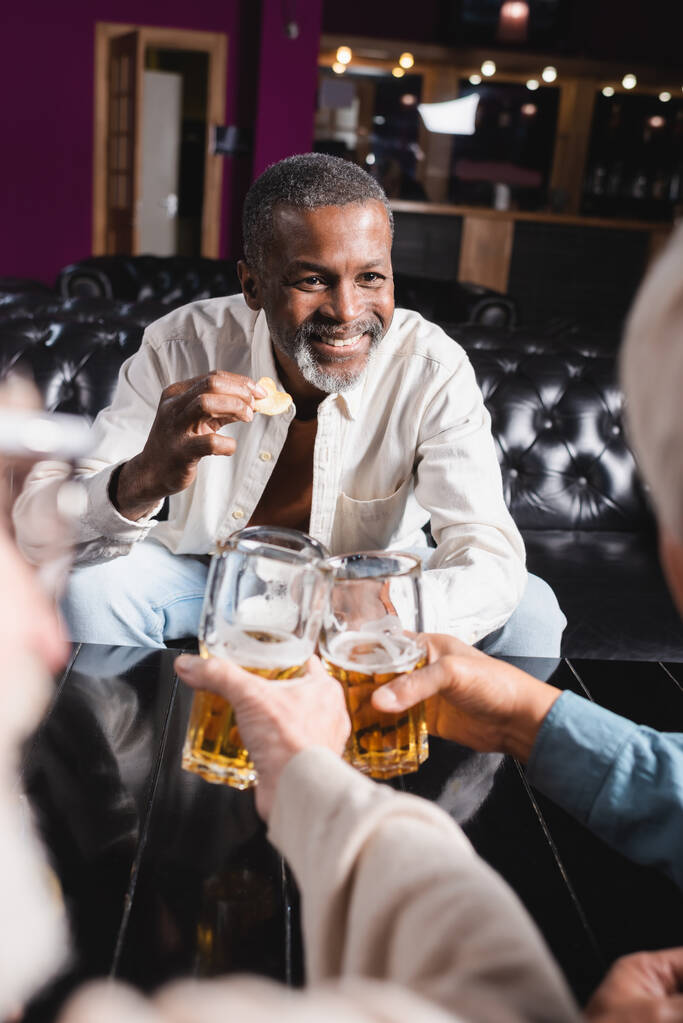 щасливий старший афроамериканський чоловік тримає чіпси під час клонування келихів пива з розмитими друзями
 - Фото, зображення