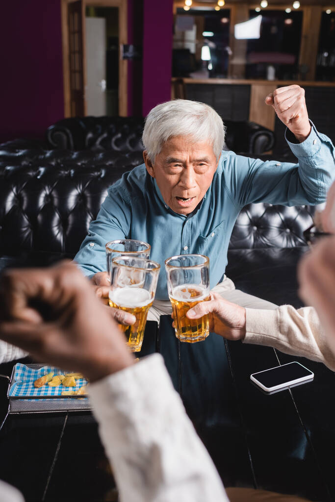 excited senior asian man showing win gesture while clinking beer glasses with blurred interracial friends - Photo, Image