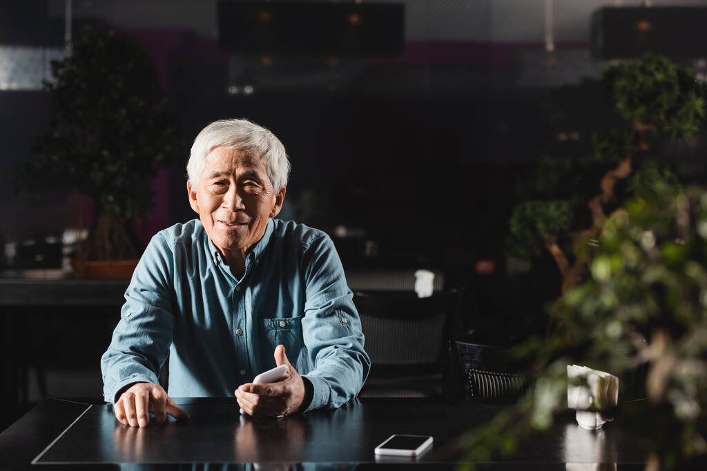 vanhempi aasialainen mies, jolla on älypuhelin ja hymyilee kameralle istuessaan kahvilassa - Valokuva, kuva