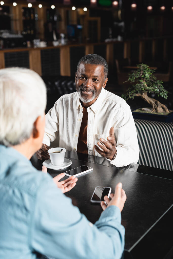 Χαρούμενος Αφροαμερικάνος που μιλάει σε θολή ηλικιωμένη φίλη κοντά σε smartphones με λευκή οθόνη στο καφέ - Φωτογραφία, εικόνα