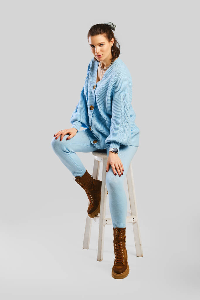 Portrait pleine longueur de belle femme en tracksuit tricoté chaud bleu clair assis sur tabouret sur fond gris - Photo, image
