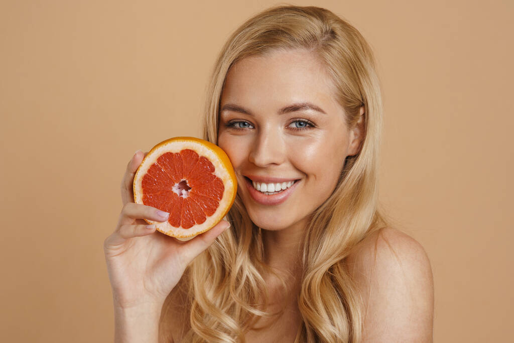Усміхнена молода блондинка топлес жінка з довгим волоссям, що стоїть над бежевим тлом, тримає розрізаний грейпфрут
 - Фото, зображення