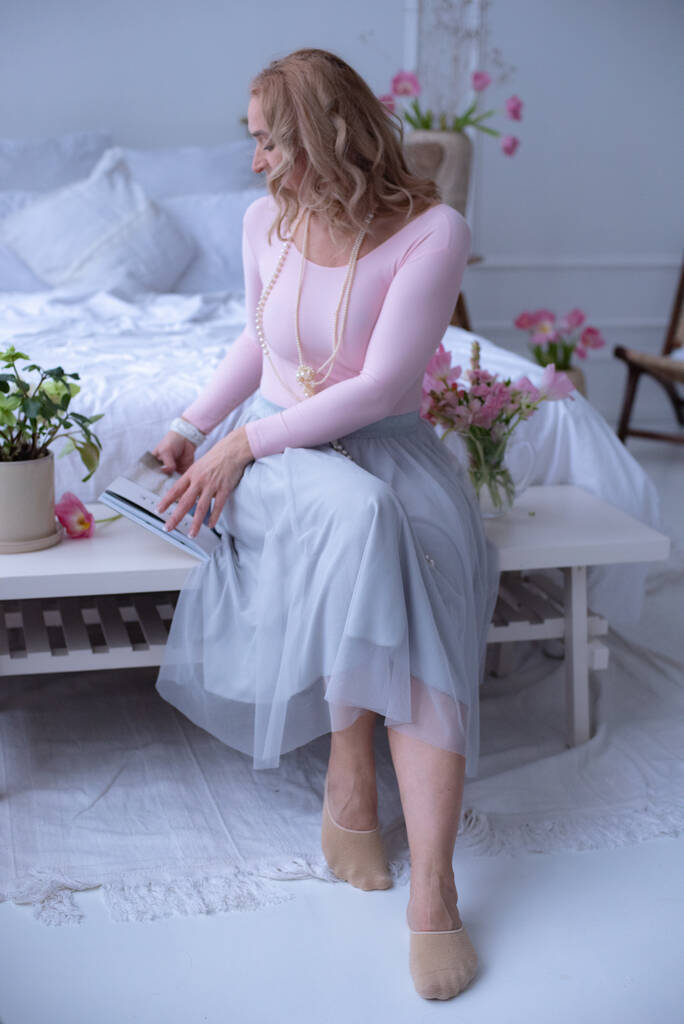 Roztomilá dívka s krátkými kudrnatými vlasy v dlouhé světle růžové sukni sedí na bílé posteli v jasném pokoji a čte si časopis. Rostliny. Dlouhé perlové korálky na krku dívky. - Fotografie, Obrázek