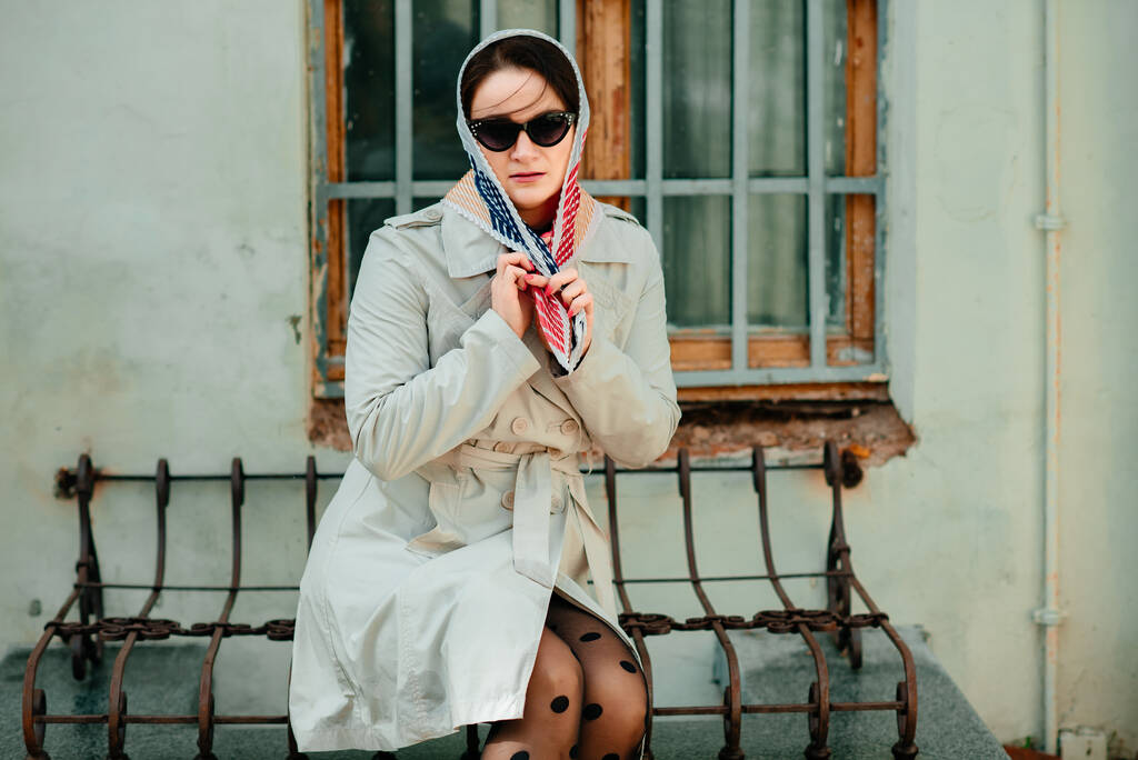 Uma menina com um lenço na cabeça em um casaco de trincheira e óculos de sol senta-se em um banco por uma velha janela de madeira na cidade. Retrato de uma menina com um lenço e óculos. - Foto, Imagem
