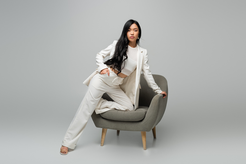 longitud completa de la joven mujer asiática apoyada en el sillón mientras posa con la mano en el bolsillo en gris - Foto, Imagen