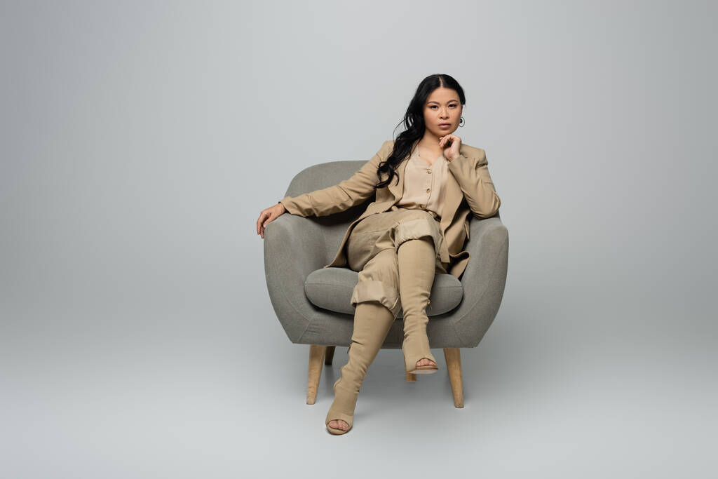 plná délka stylové asijské ženy sedí v křesle na šedé - Fotografie, Obrázek