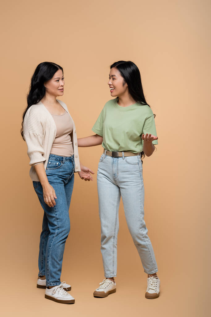 full length of happy asian mother in jeans looking at daughter on beige  - Photo, Image