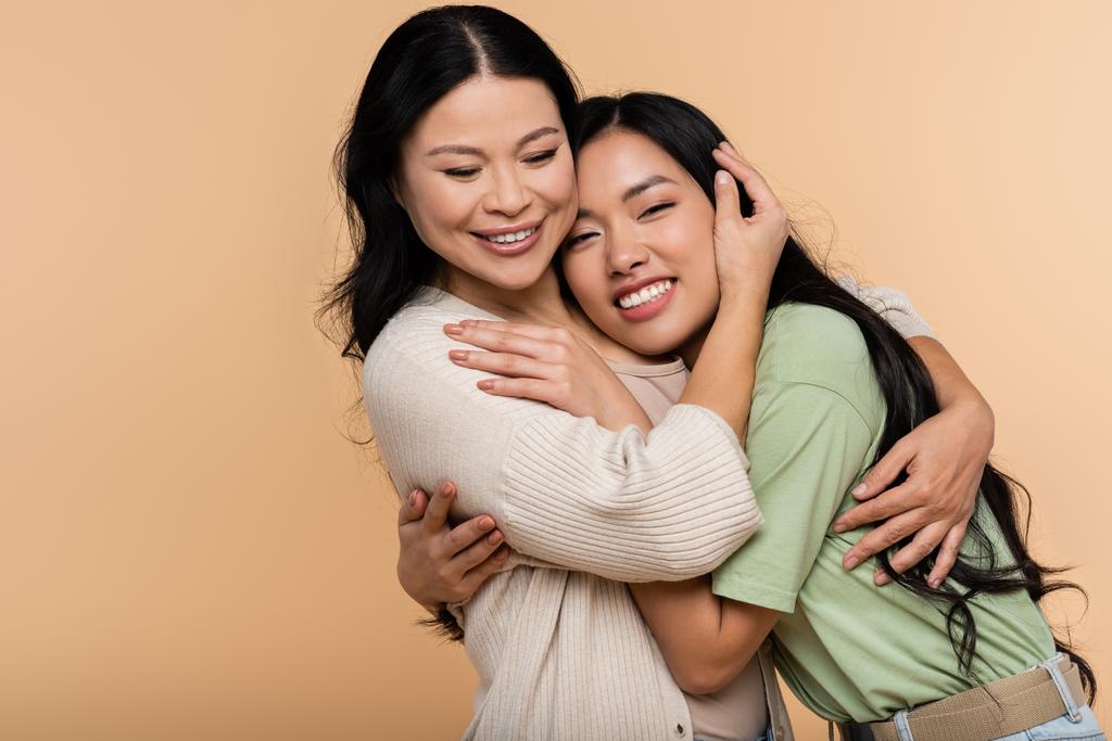 vrolijk aziatisch moeder en dochter knuffelen geïsoleerd op beige - Foto, afbeelding