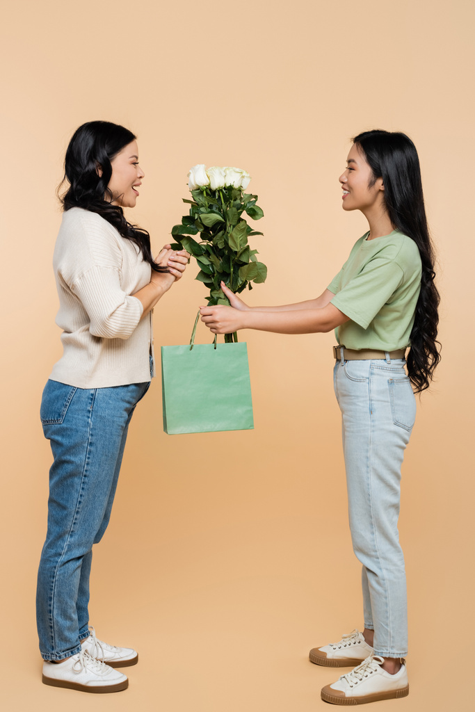 full length of happy asian daughter giving paper bag and flowers to mother on beige - Photo, Image