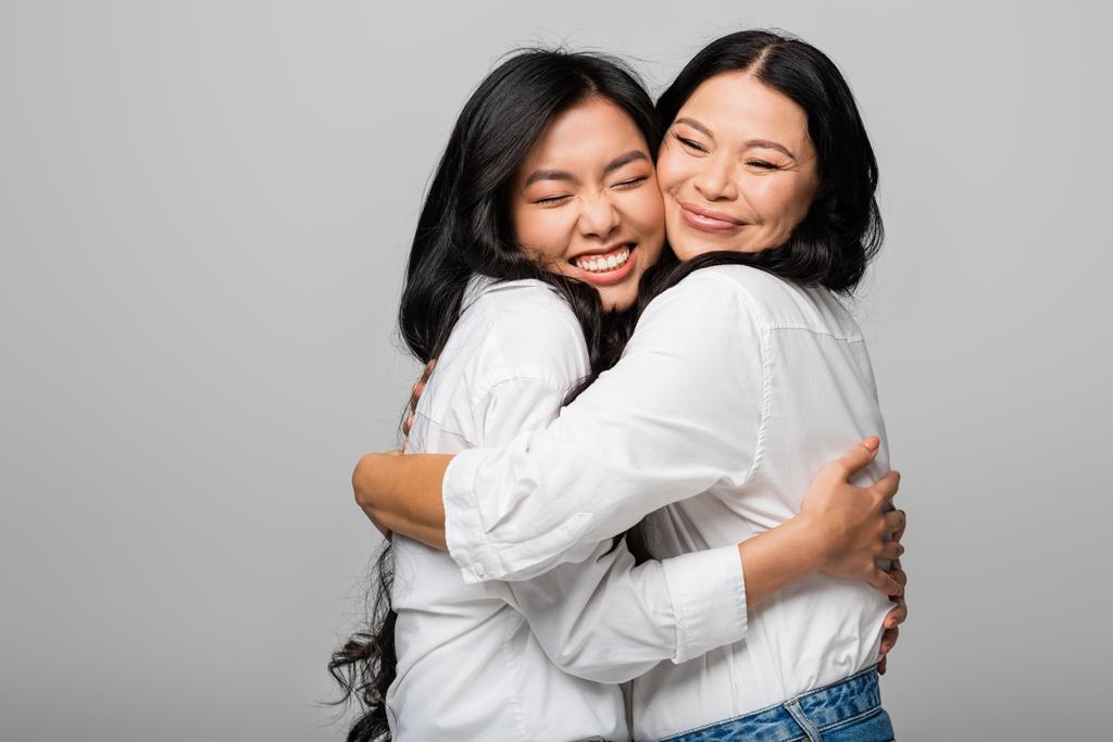 complacido asiático madre y hija en blanco camisas abrazo aislado en gris - Foto, imagen