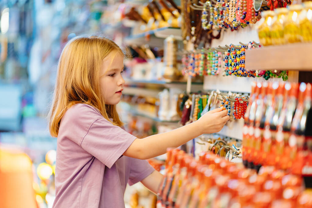 ένα αστείο κοριτσάκι κοιτάζει με ενδιαφέρον σε κοσμήματα και σουβενίρ στο κατάστημα.  - Φωτογραφία, εικόνα