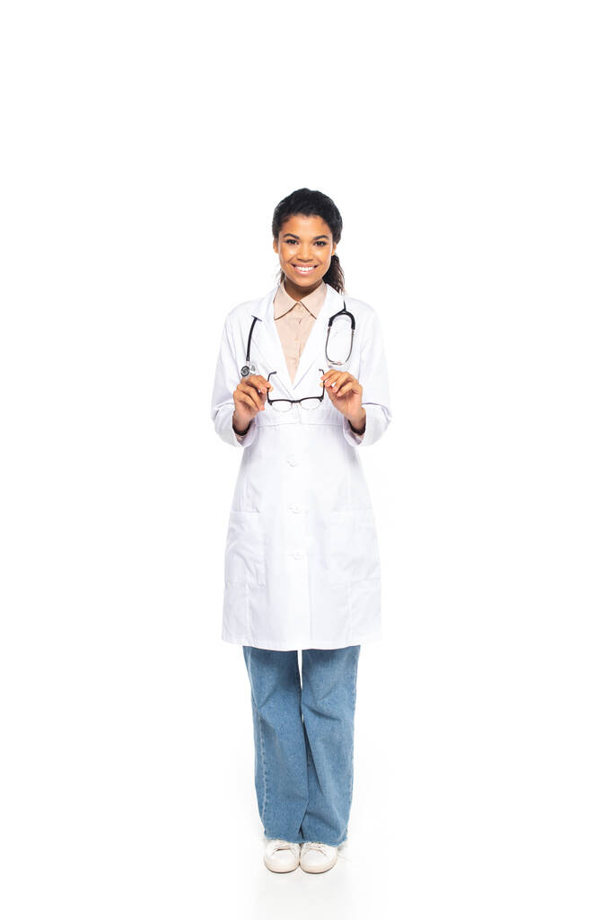 Longitud completa de sonriente médico afroamericano sosteniendo anteojos sobre fondo blanco - Foto, imagen