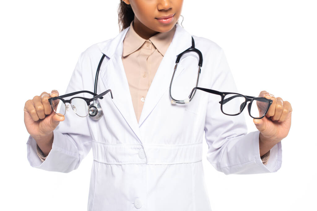 Обрізаний вид афроамериканського окуліста в білому пальто і стетоскоп, що тримає окуляри ізольовані на білому
  - Фото, зображення