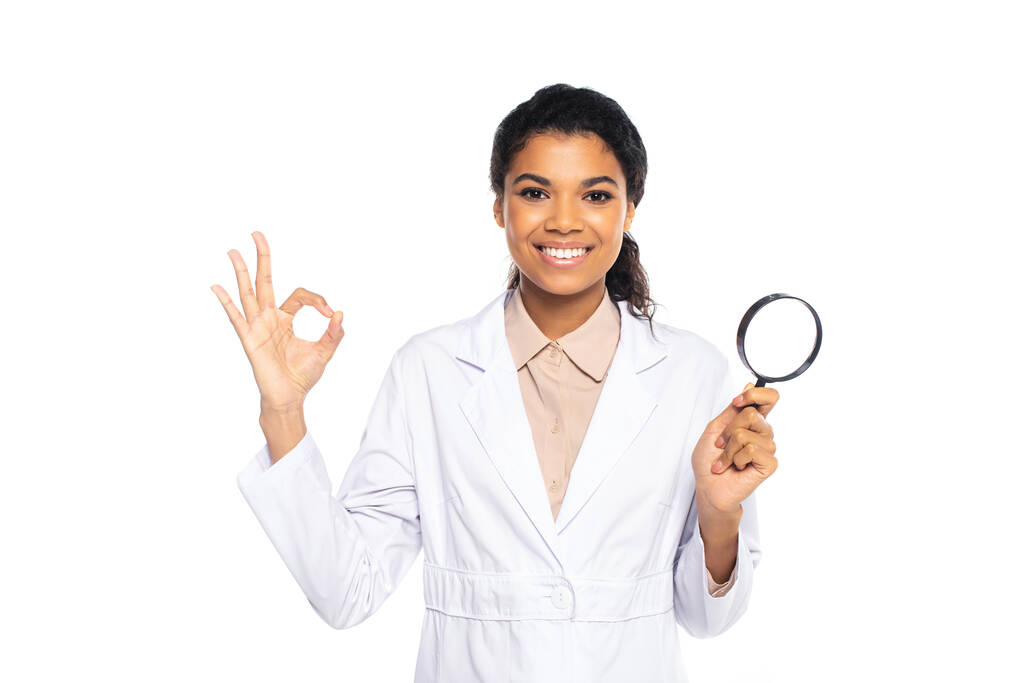 African american doctor in while coat holding magnifying glass and showing ok gesture isolated on white  - Photo, Image